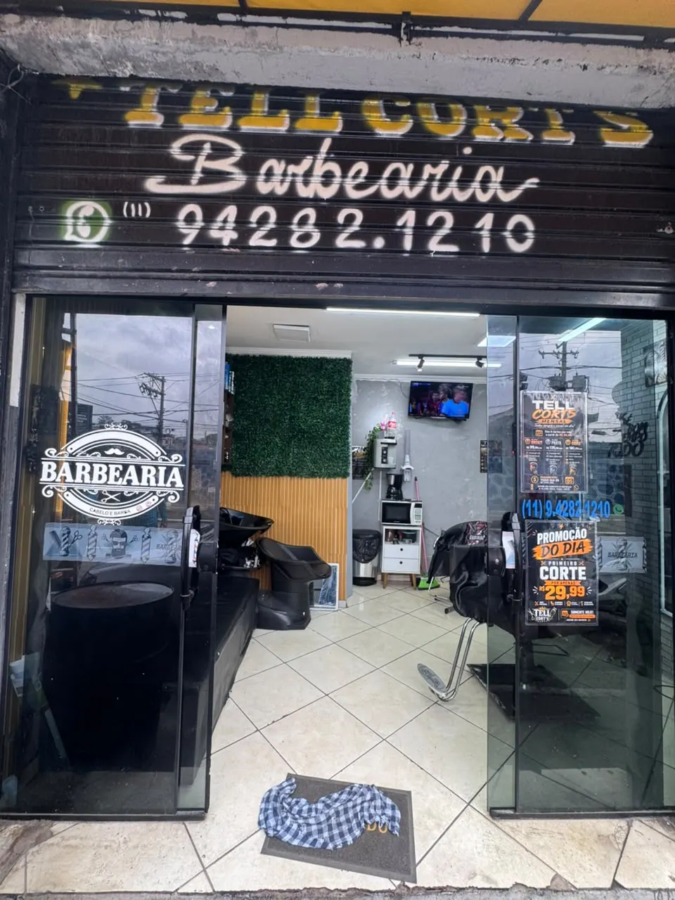 Fachada da barbearia na Estrada Turística do Jaraguá, com o nome pintado na porta de aço e o telefone